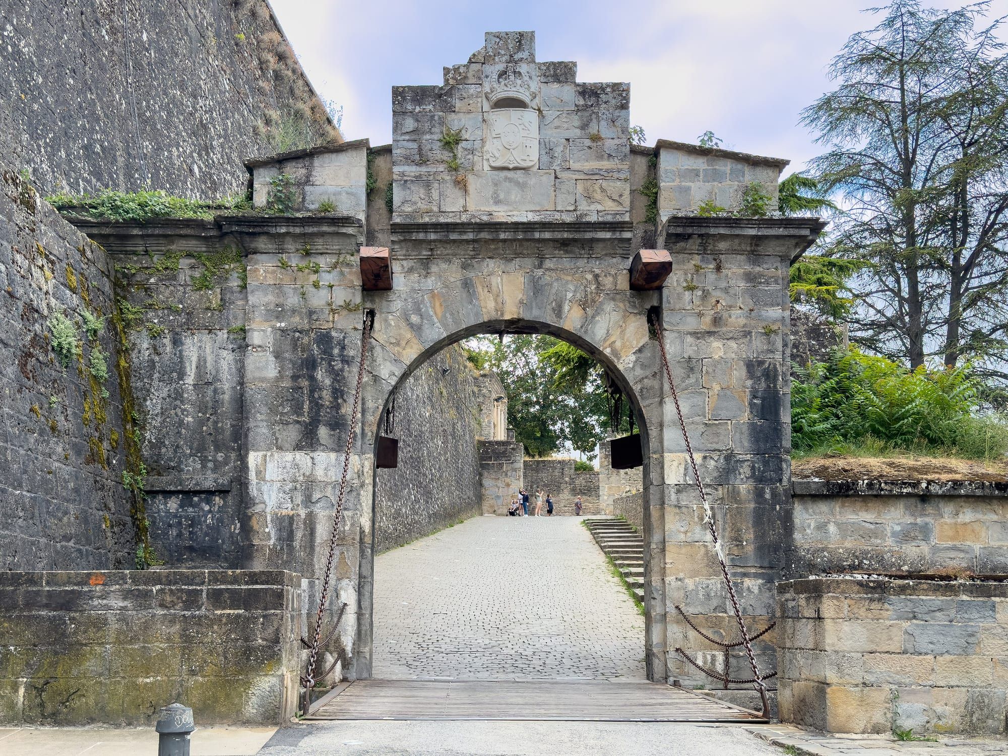 The Portal de Francia, the gate in Pamplona's Renaissance walls through which the Camino Francés enters the old town: a weathered sandstone gateway topped by a stepped crest carved with a crowned coat of arms, its round arch spanned by a wooden drawbridge deck hung from iron chains and counterweights, opens onto a cobbled ramp climbing between high ramparts towards a few tiny distant visitors, with the massive moss-streaked bastion wall on the left and ferns and tall cedars on the right under a pale summer sky