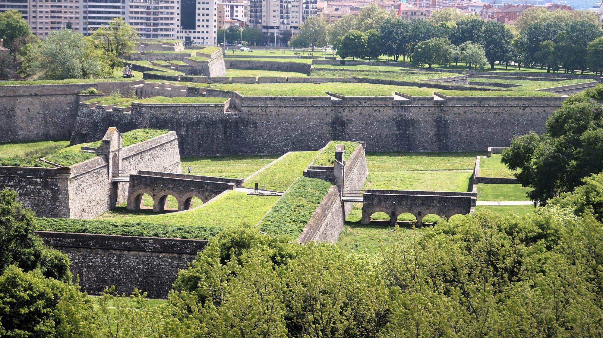 The outer defences of Pamplona's Ciudadela, the star-shaped sixteenth-century fortress, seen from above in spring: grass-topped stone ravelins and counterguards step down in sharp angular terraces to dry moats crossed by low arched stone bridges, framed by leafy treetops of the Vuelta del Castillo park in the foreground, with more ramparts, a line of trees and apartment blocks in the distance