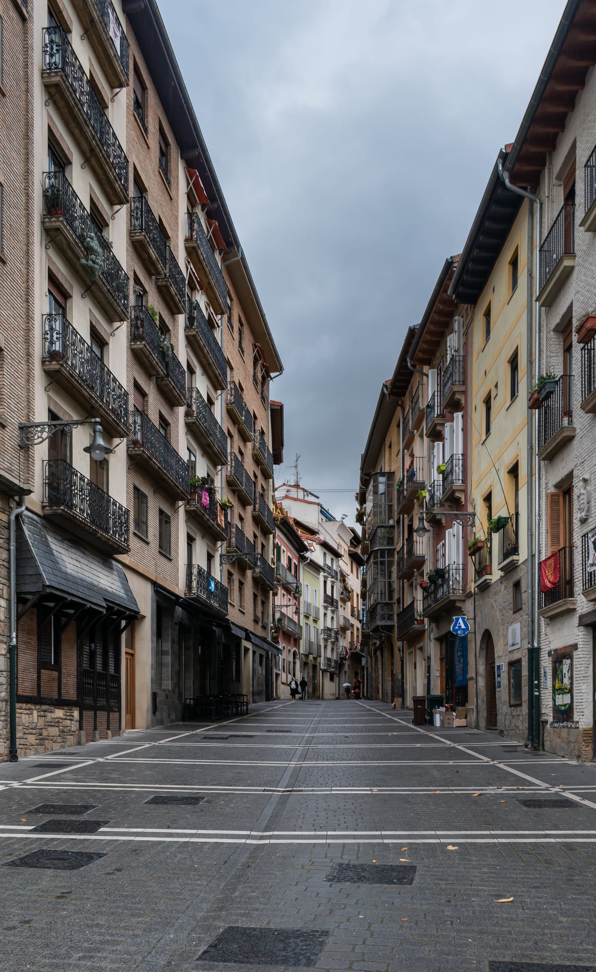 Calle del Carmen, the first street of Pamplona's old town that pilgrims walk after the Portal de Francia, almost empty on an overcast day: a straight stone-paved street with pale guide lines runs between tall houses with iron balconies, flower pots and wall lanterns, a small blue pilgrim-hostel sign with a scallop shell hangs on the right, and a few distant figures walk at the far end under a heavy grey sky