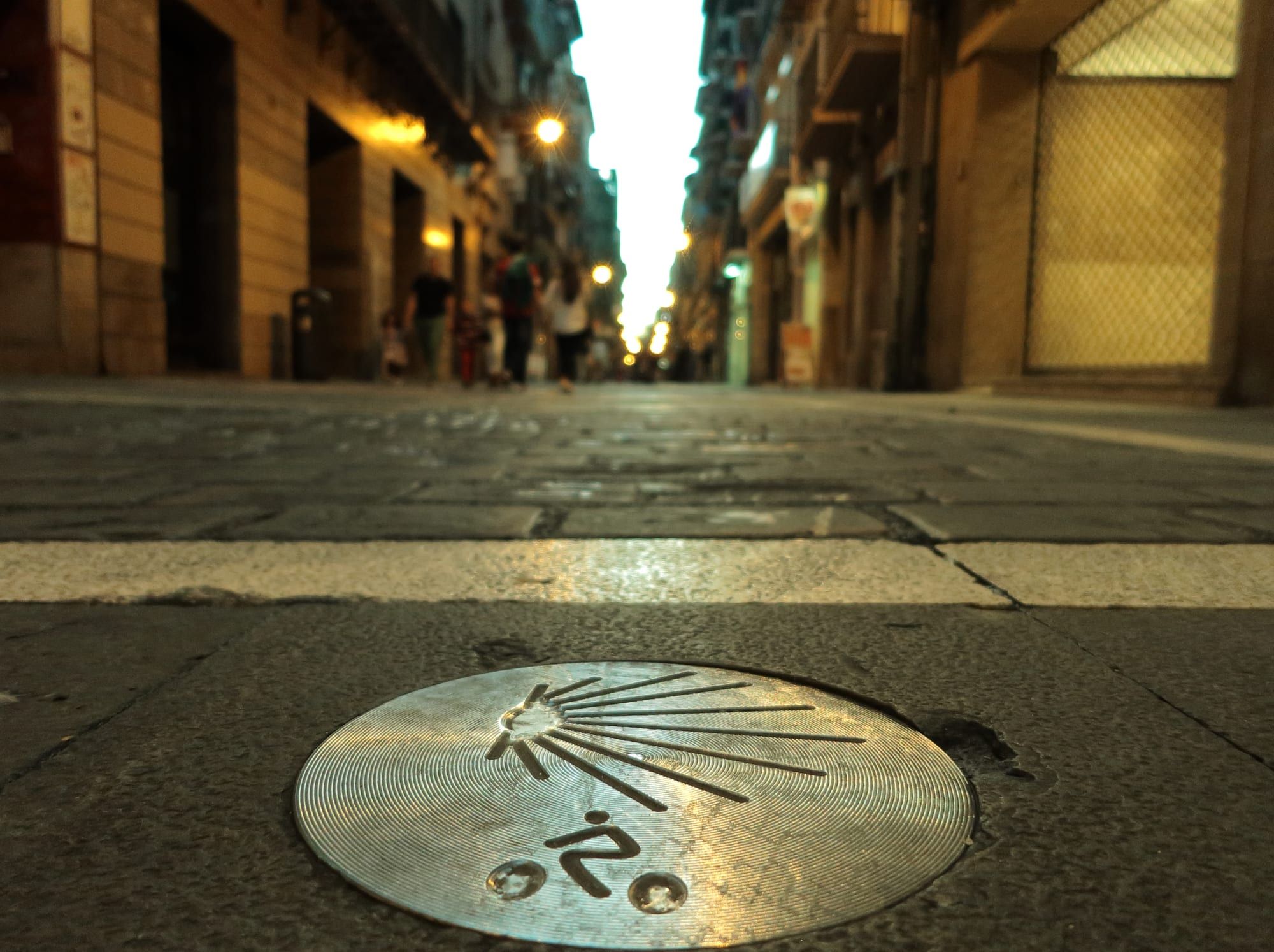 A Camino de Santiago marker set into the paving of a street in Pamplona's old town at dusk: a round polished metal disc engraved with a scallop shell and a small stylised emblem gleams sharp in the foreground across a white stripe in the stone, while the narrow street beyond, lined with tall houses and lit by warm street lamps, fades out of focus with a few blurred passers-by