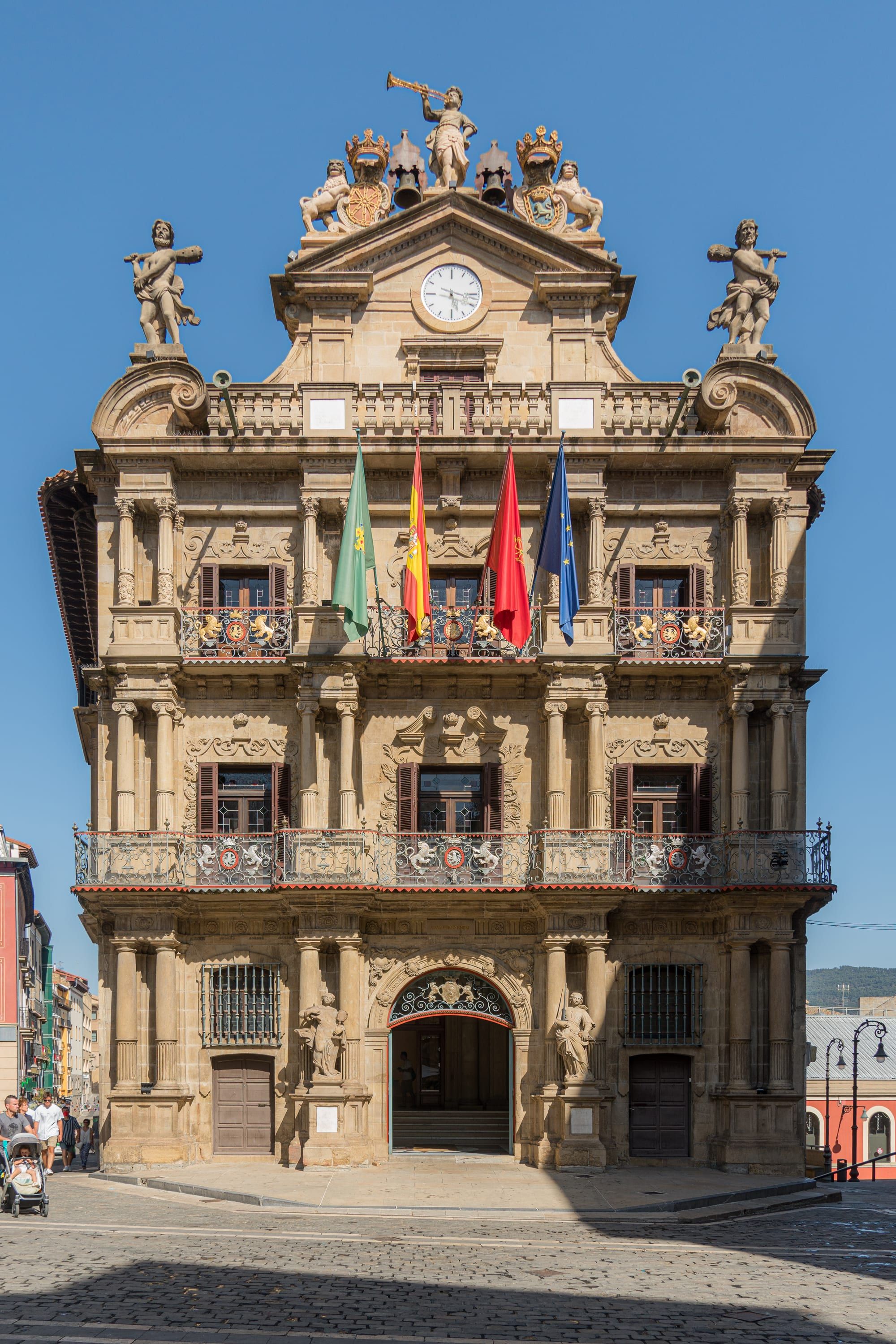 The Casa Consistorial, Pamplona's eighteenth-century town hall, seen head-on under a deep blue summer sky: a tall Baroque façade of golden stone rising in three orders of columns, with wrought-iron balconies hung with painted coats of arms, four flags on the main balcony, a clock in the pediment and allegorical statues and a trumpeting figure on the roofline, a cobbled square in front and a few small passers-by at the far left edge